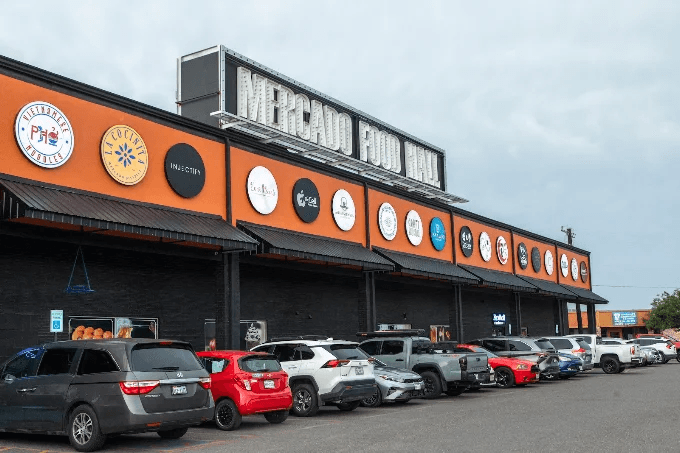 Exterior of Mercado Food Hall & Shops in McAllen: a long orange facade with the market's sign on the roof and the tenants' logos along the front.
